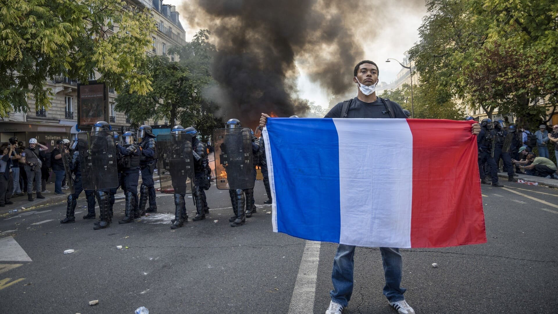 À Paris, un manifestant tient un drapeau français devant un cordon de policiers casqués, tandis qu’un incendie fait rage derrière eux. © pmvfoto / Shutterstock