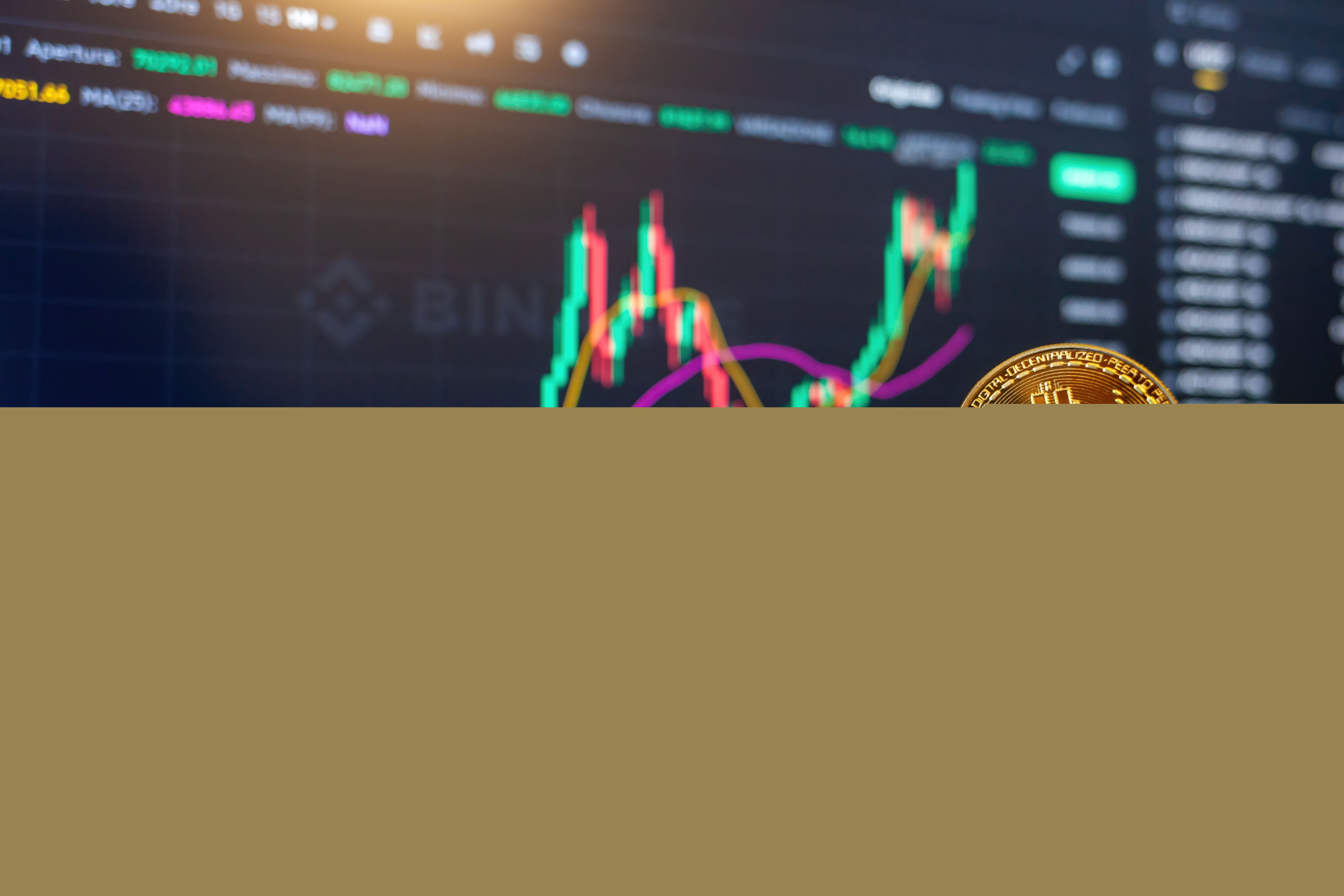 Une pièce de Bitcoin tenu devant un écran de trading © Shutterstock