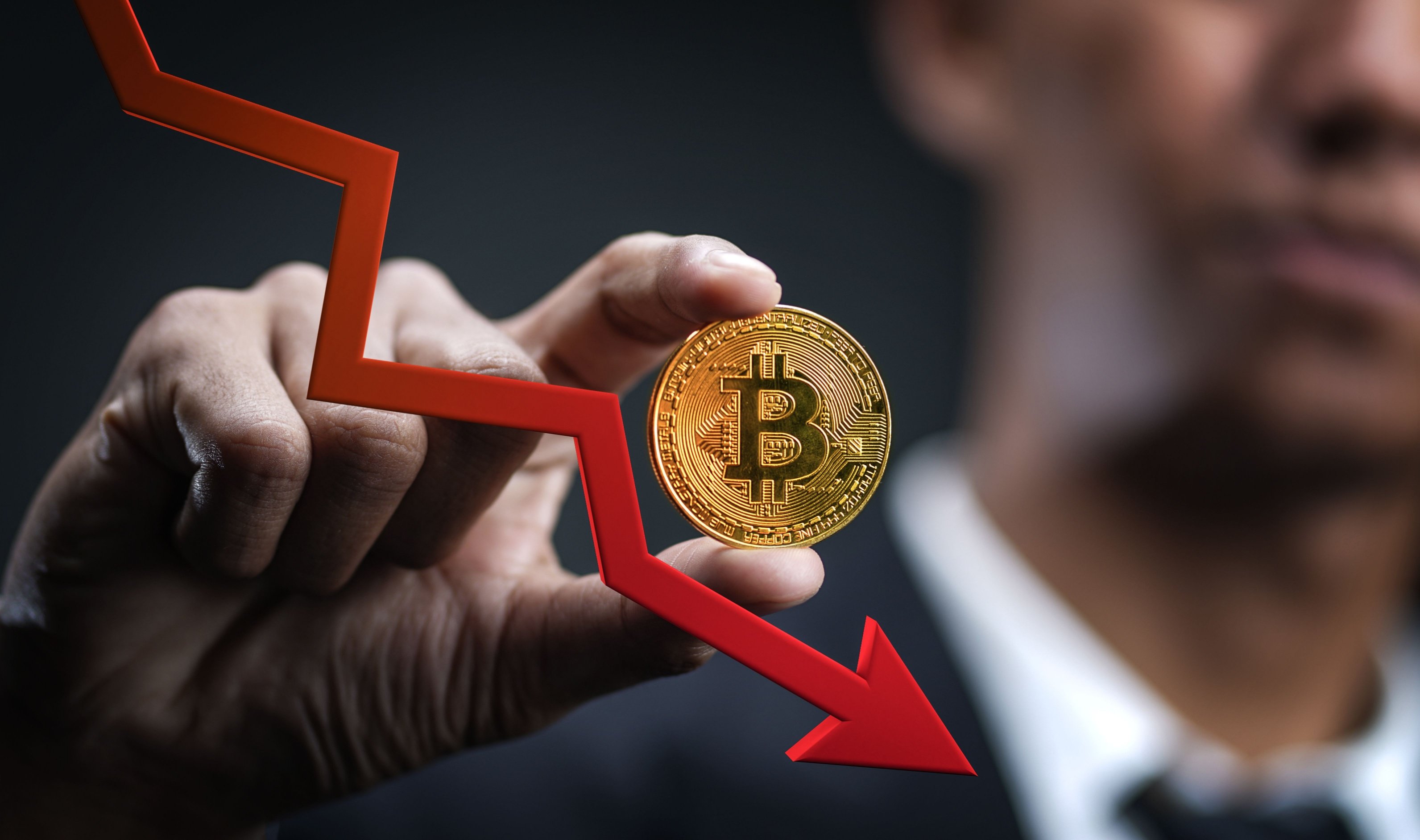 Un homme tient une pièce Bitcoin alors qu'une flèche rouge indique la chute de son cours © Shutterstock