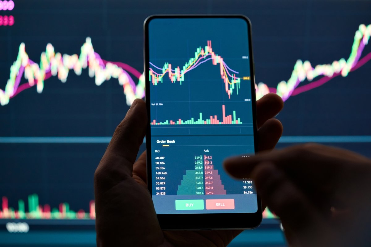 Un écran de smartphone affichant le cours d'une cryptomonnaie, devant un écran similaire plus grand© Ground Picture / Shutterstock