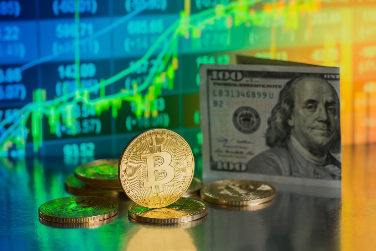 Une pièce estampillée Bitcoin et un billet de cent dollars font face à un écran affichant des cours boursiers © Shutterstock